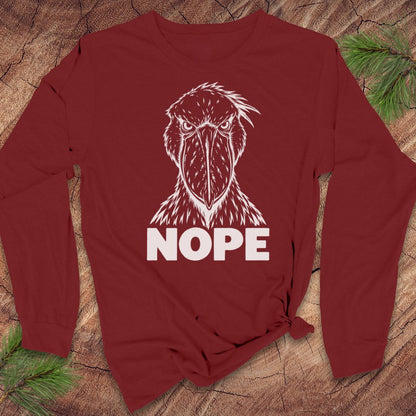 Cardinal red long-sleeve t-shirt with a white line-art Shoebill staring straight ahead above the word NOPE, laid flat on a rustic wood background with pine branches.