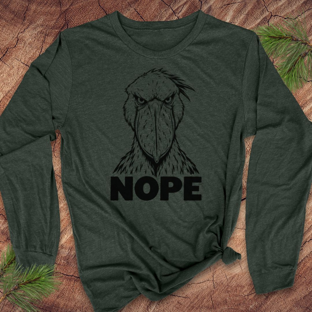 Heather forest green long-sleeve t-shirt with a black line-art Shoebill staring straight ahead above the word NOPE, laid flat on a rustic wood background with pine branches.