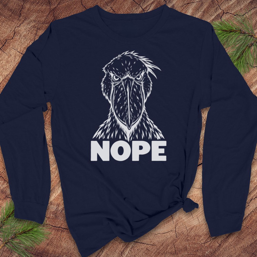 Navy long-sleeve t-shirt with a white line-art Shoebill staring straight ahead above the word NOPE, laid flat on a rustic wood background with pine branches.