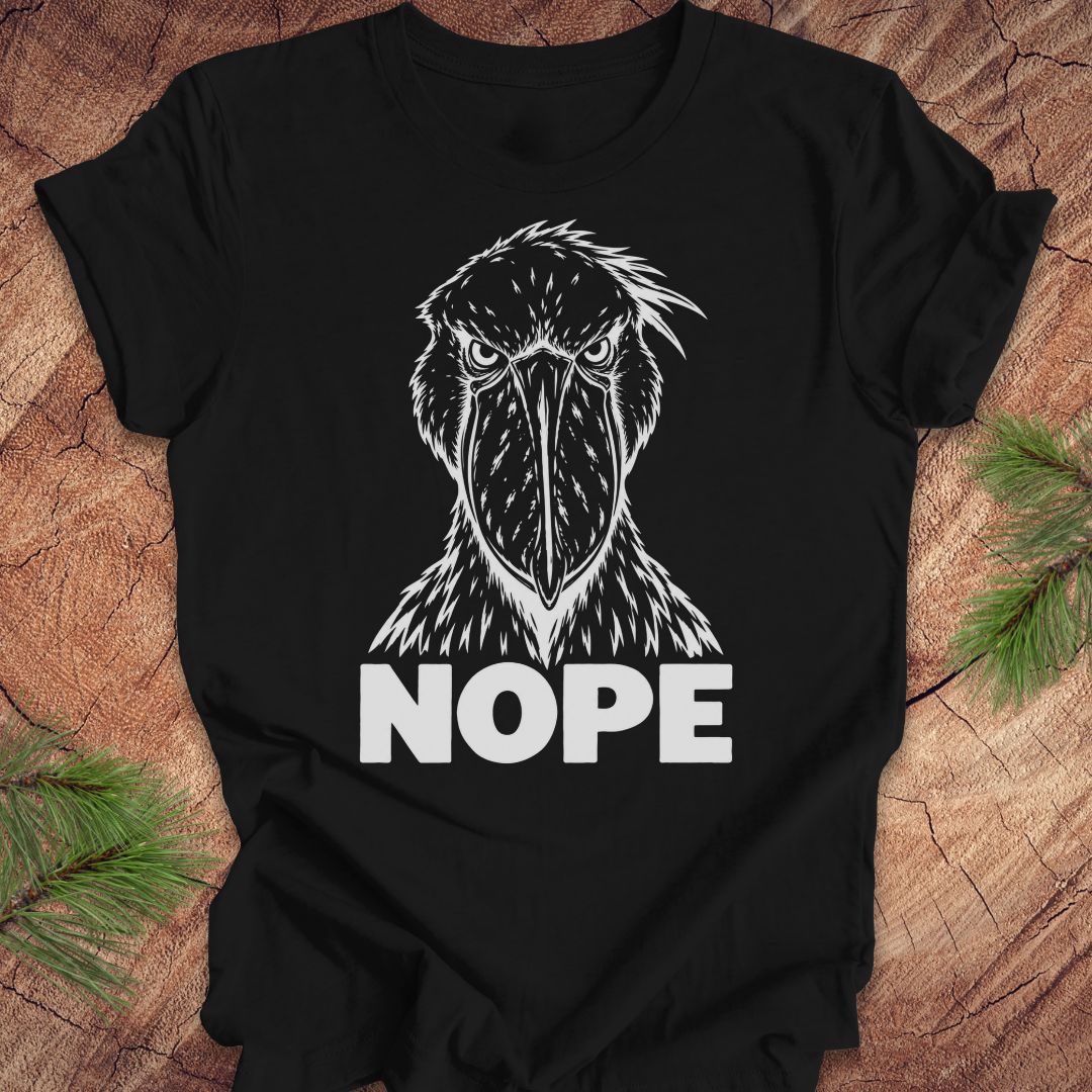 Black t-shirt with a white line-art Shoebill staring straight ahead above the word NOPE, laid flat on a rustic wood background with pine branches.