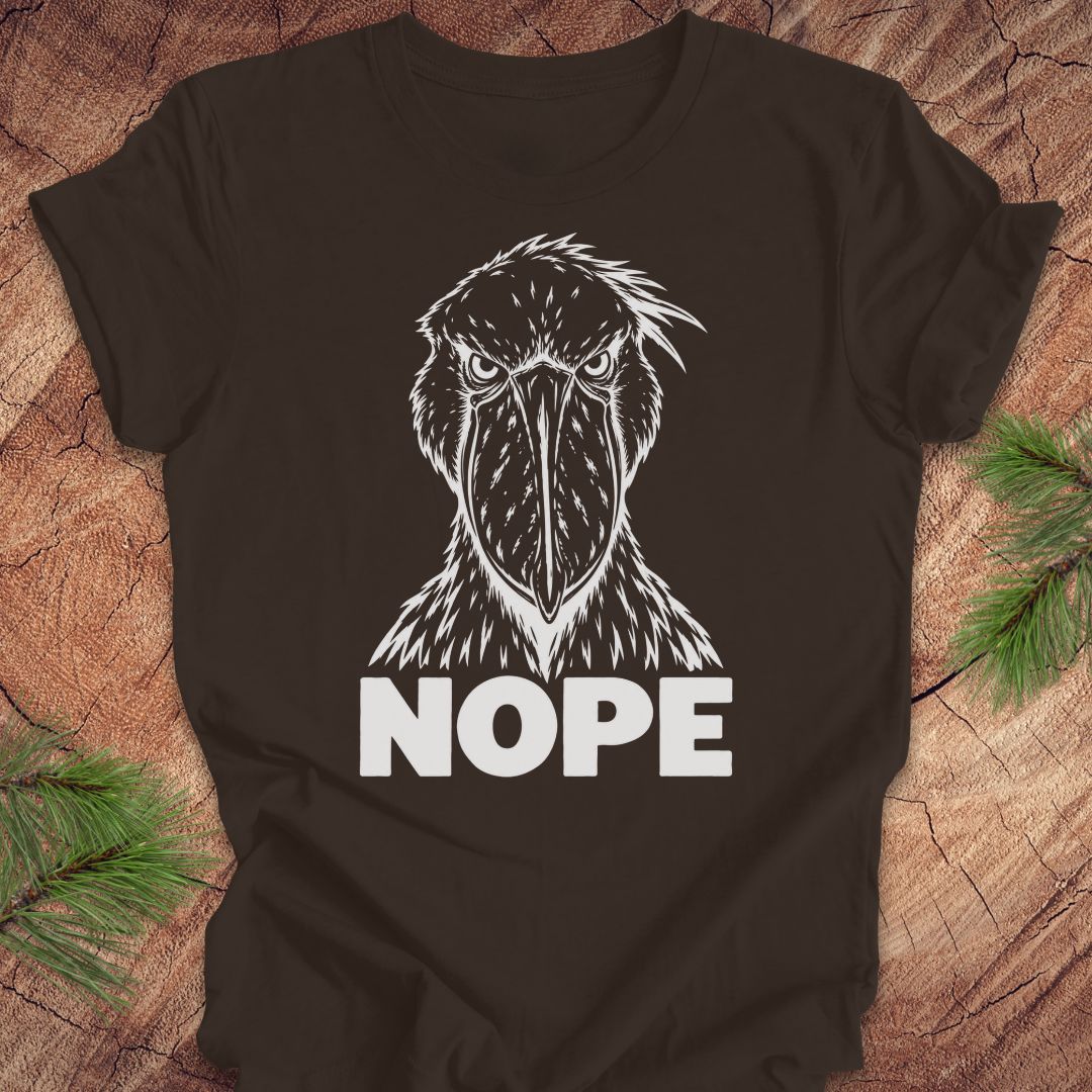 Brown t-shirt with a white line-art Shoebill staring straight ahead above the word NOPE, laid flat on a rustic wood background with pine branches.