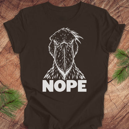 Brown t-shirt with a white line-art Shoebill staring straight ahead above the word NOPE, laid flat on a rustic wood background with pine branches.