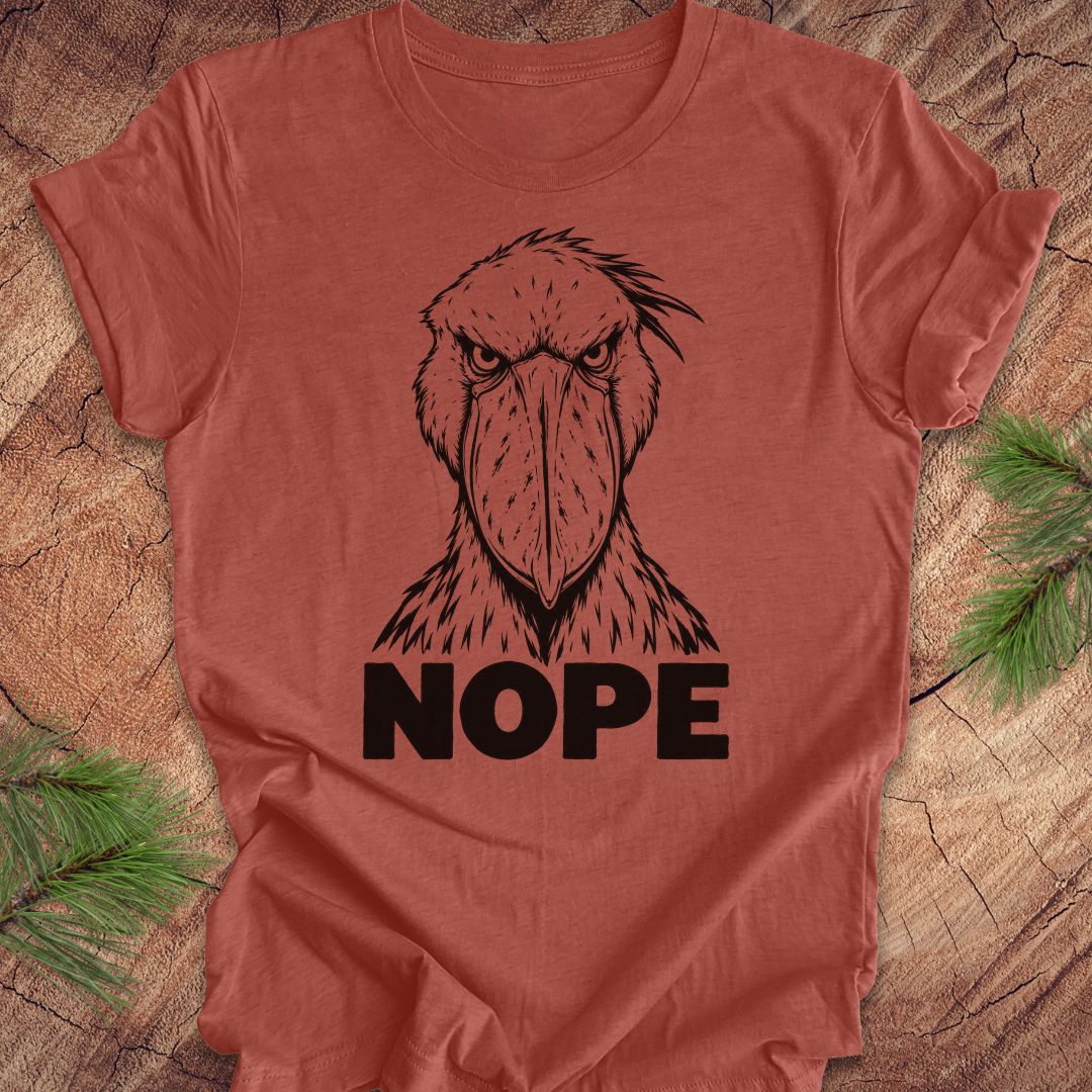 Heather clay rust t-shirt with a white line-art Shoebill staring straight ahead above the word NOPE, laid flat on a rustic wood background with pine branches.