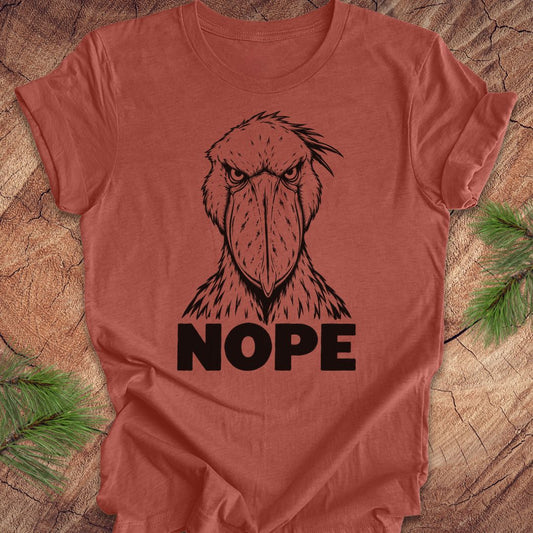 Heather clay rust t-shirt with a white line-art Shoebill staring straight ahead above the word NOPE, laid flat on a rustic wood background with pine branches.