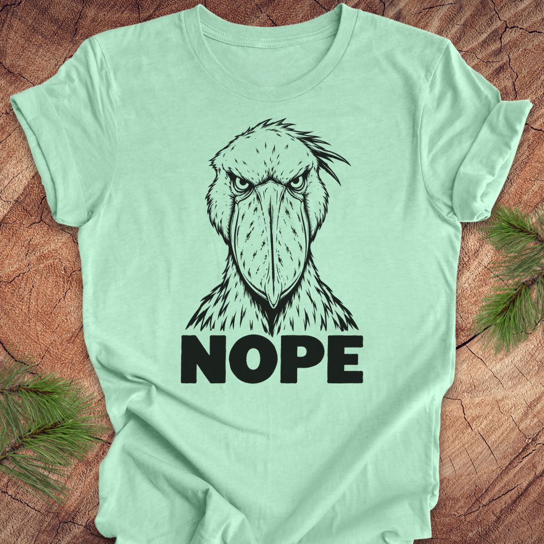 Heather mint green t-shirt with a white line-art Shoebill staring straight ahead above the word NOPE, laid flat on a rustic wood background with pine branches.