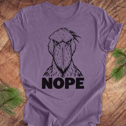 Heather purple t-shirt with a white line-art Shoebill staring straight ahead above the word NOPE, laid flat on a rustic wood background with pine branches.
