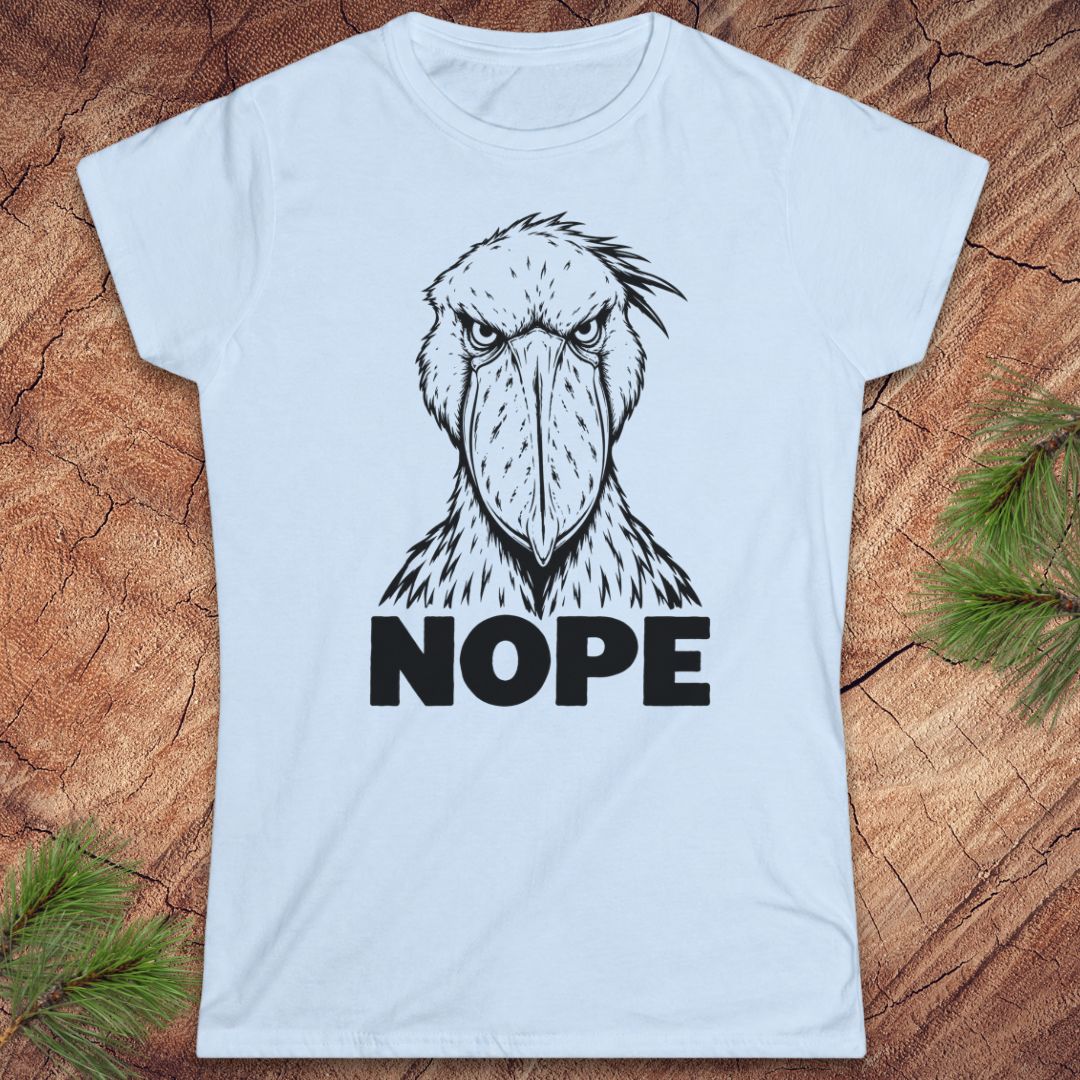 Light blue women's t-shirt with a black line-art Shoebill staring straight ahead above the word NOPE, laid flat on a rustic wood background with pine branches.