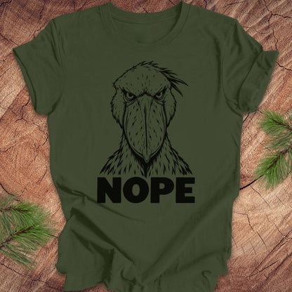 Military green t-shirt with a white line-art Shoebill staring straight ahead above the word NOPE, laid flat on a rustic wood background with pine branches.
