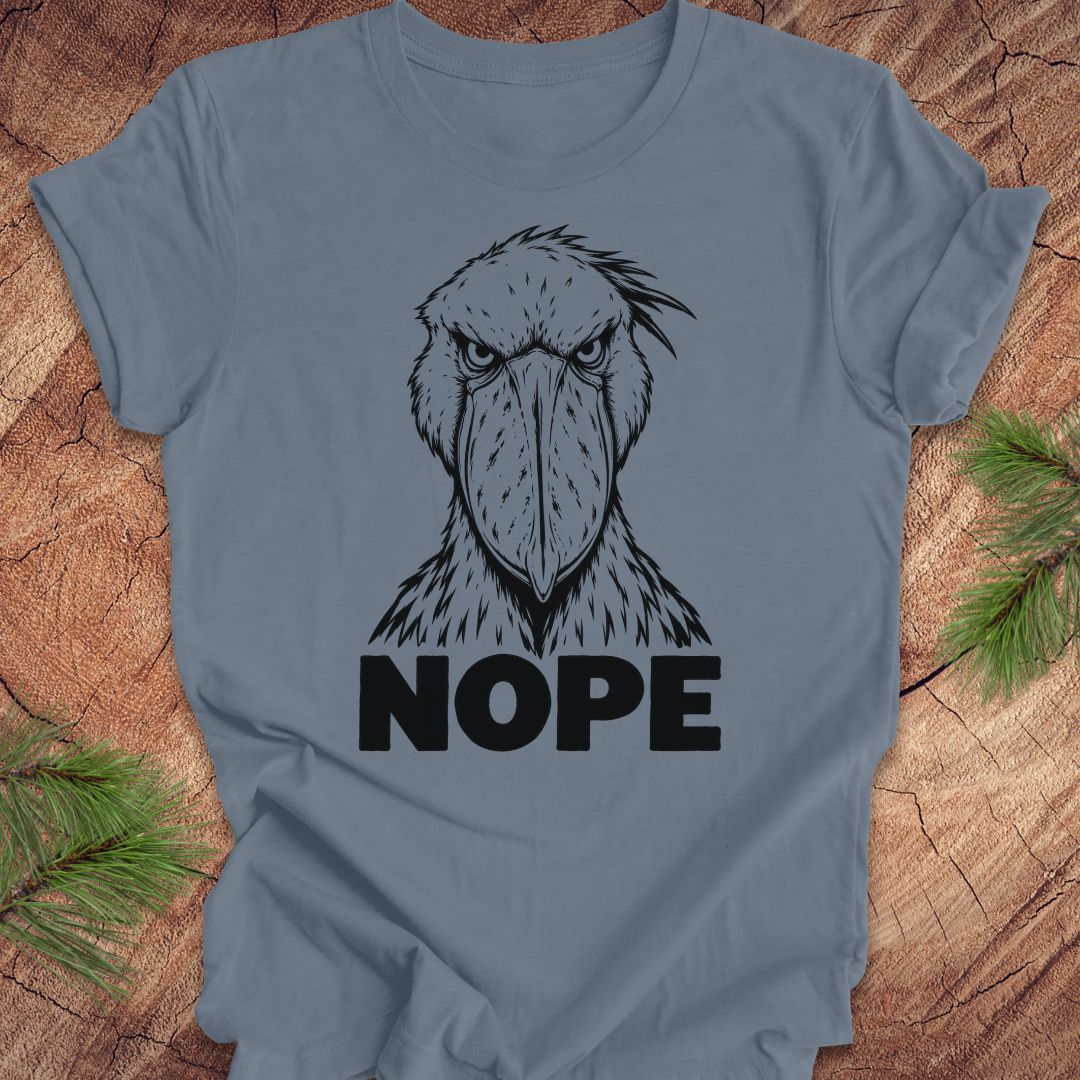 Steel blue t-shirt with a white line-art Shoebill staring straight ahead above the word NOPE, laid flat on a rustic wood background with pine branches.