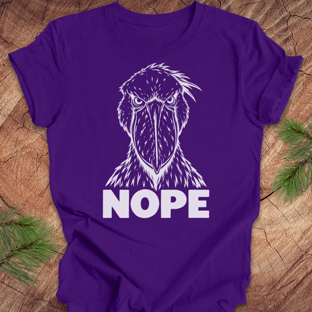 Purple t-shirt with a white line-art Shoebill staring straight ahead above the word NOPE, laid flat on a rustic wood background with pine branches.