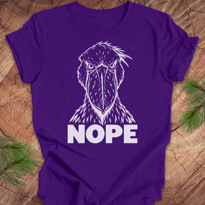 Purple t-shirt with a white line-art Shoebill staring straight ahead above the word NOPE, laid flat on a rustic wood background with pine branches.