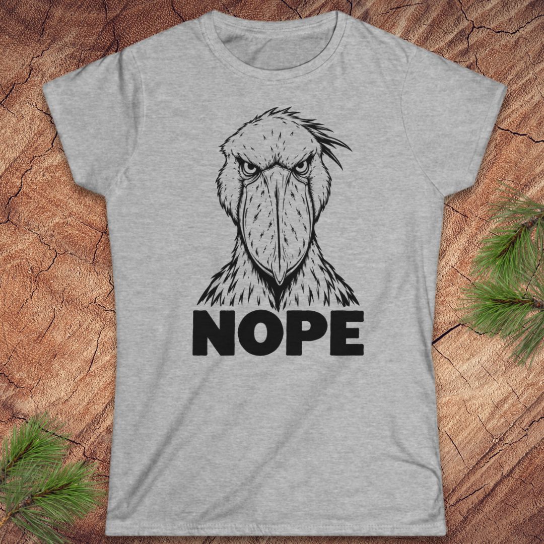 Sport grey women's t-shirt with a black line-art Shoebill staring straight ahead above the word NOPE, laid flat on a rustic wood background with pine branches.