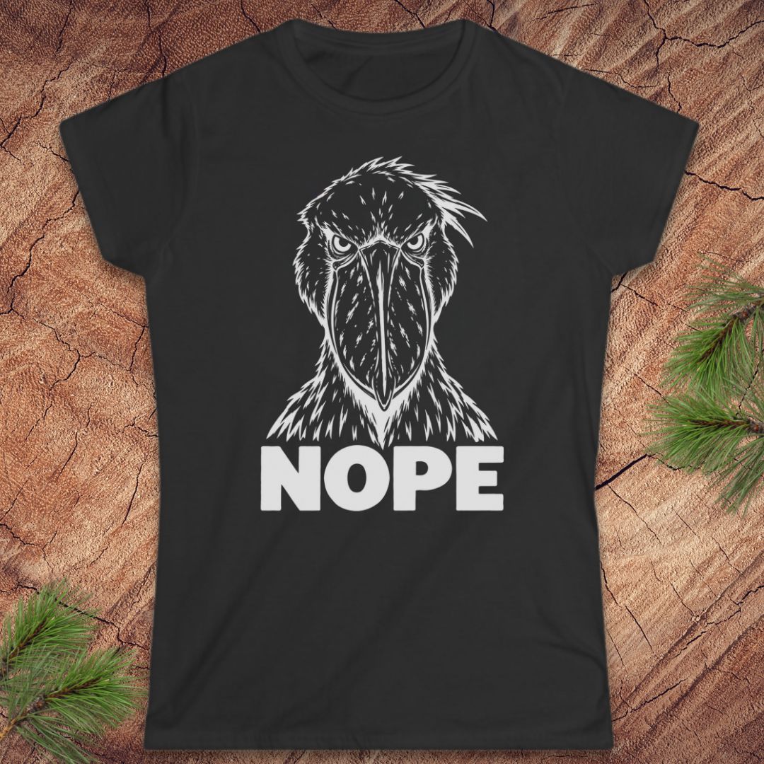 Black women's t-shirt with a white line-art Shoebill staring straight ahead above the word NOPE, laid flat on a rustic wood background with pine branches.