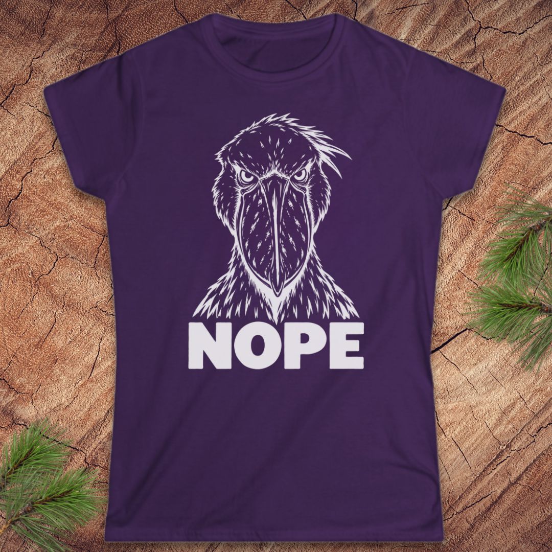 Purple women's t-shirt with a white line-art Shoebill staring straight ahead above the word NOPE, laid flat on a rustic wood background with pine branches.