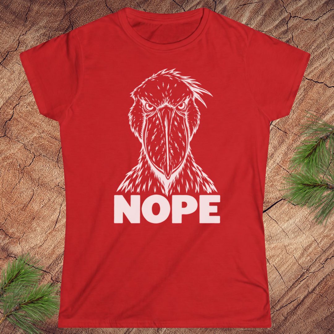 Red women's t-shirt with a white line-art Shoebill staring straight ahead above the word NOPE, laid flat on a rustic wood background with pine branches.