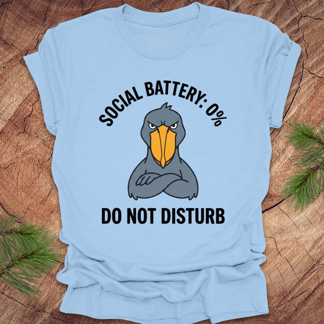Light blue t-shirt with a cartoon shoebill stork graphic and text 'social battery 0%. Do not disturb' on a wooden surface.