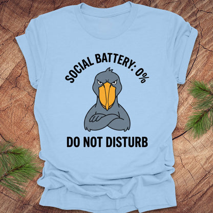 Light blue t-shirt with a cartoon shoebill stork graphic and text 'social battery 0%. Do not disturb' on a wooden surface.