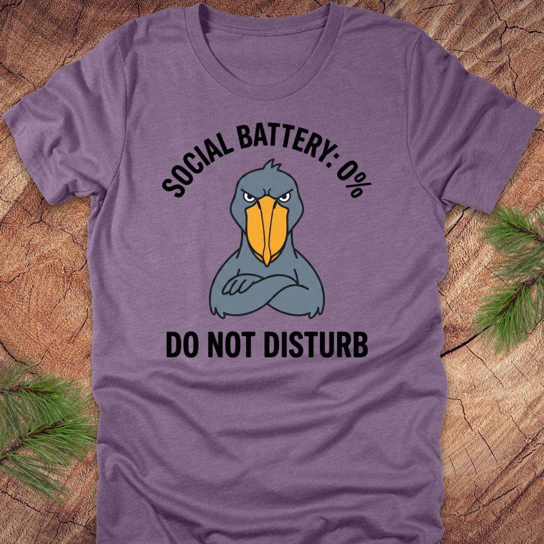 Purple t-shirt with a cartoon shoebill stork graphic and text 'social battery 0%. Do not disturb' on a wooden surface.