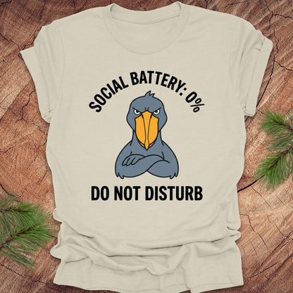 Beige t-shirt with a cartoon shoebill stork graphic and text 'social battery 0%. Do not disturb' on a wooden surface.