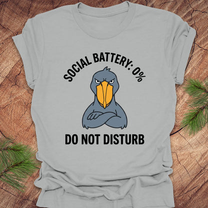 Silver t-shirt with a cartoon shoebill stork graphic and text 'social battery 0%. Do not disturb' on a wooden surface.
