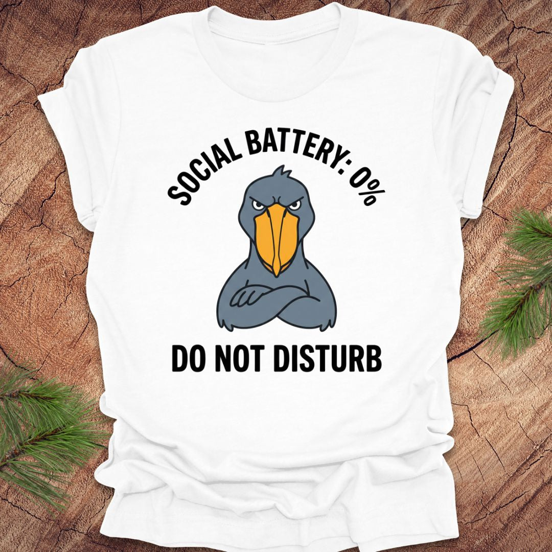 White t-shirt with a cartoon shoebill stork graphic and text 'social battery 0%. Do not disturb' on a wooden surface.