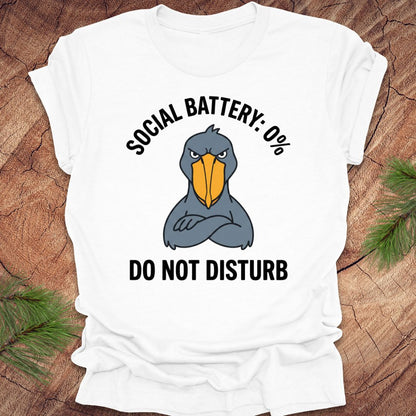 White t-shirt with a cartoon shoebill stork graphic and text 'social battery 0%. Do not disturb' on a wooden surface.