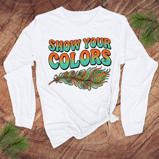 White long-sleeve shirt with 'Show Your Colors' text and peacock feather design on a wooden surface.