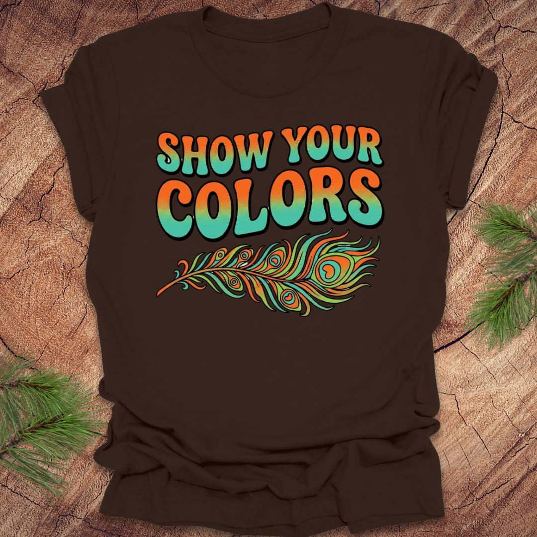 Brown t-shirt with 'Show Your Colors' text and peacock feather design on a wooden surface.