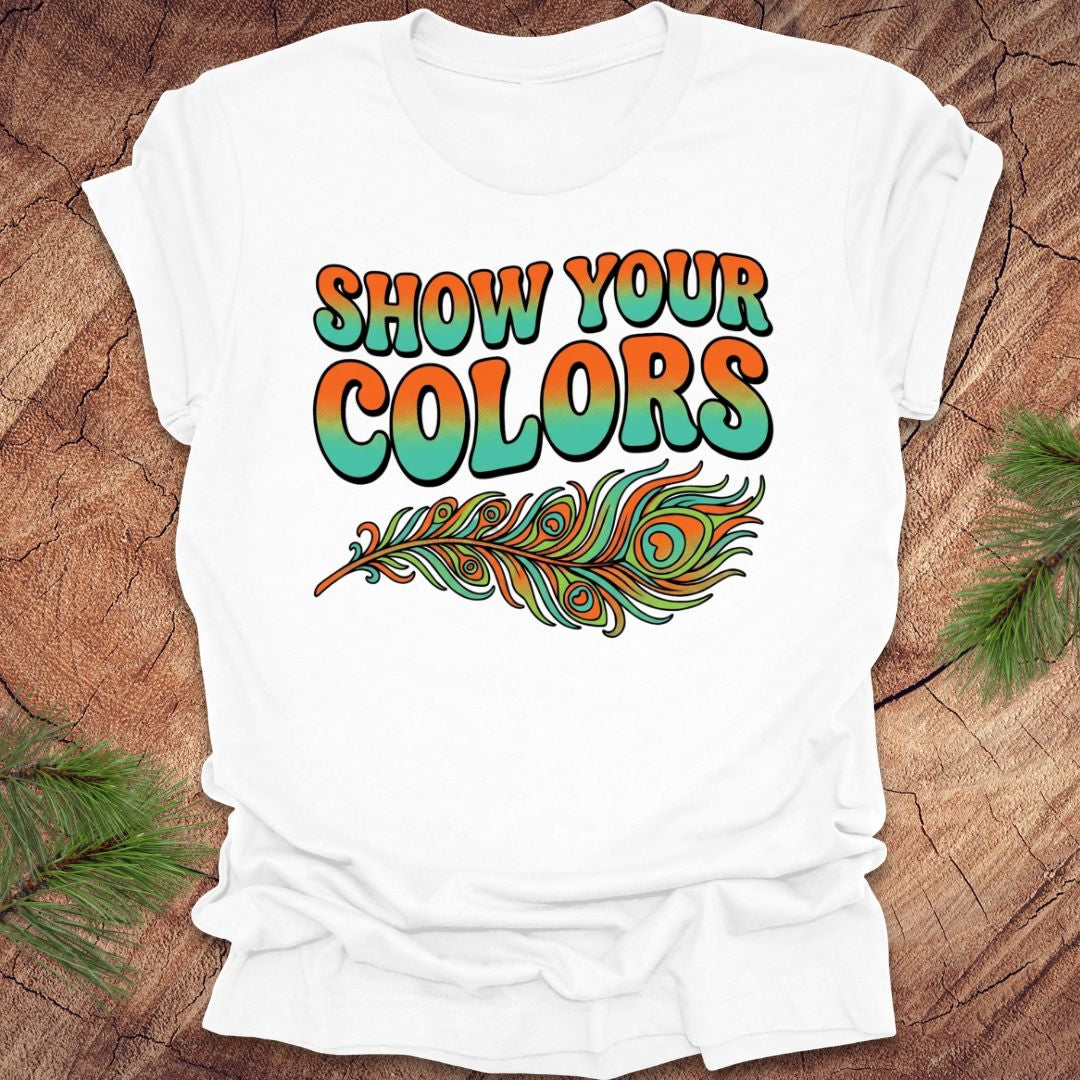White t-shirt with 'Show Your Colors' text and peacock feather design on a wooden surface.