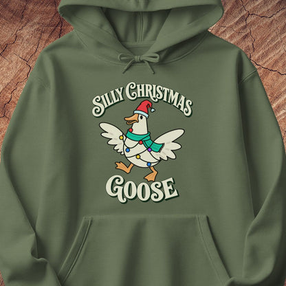 Military green hoodie with a goose in a Santa hat and teal scarf, wrapped in colorful Christmas lights under retro Silly Christmas Goose lettering, laid flat on a rustic wood background.