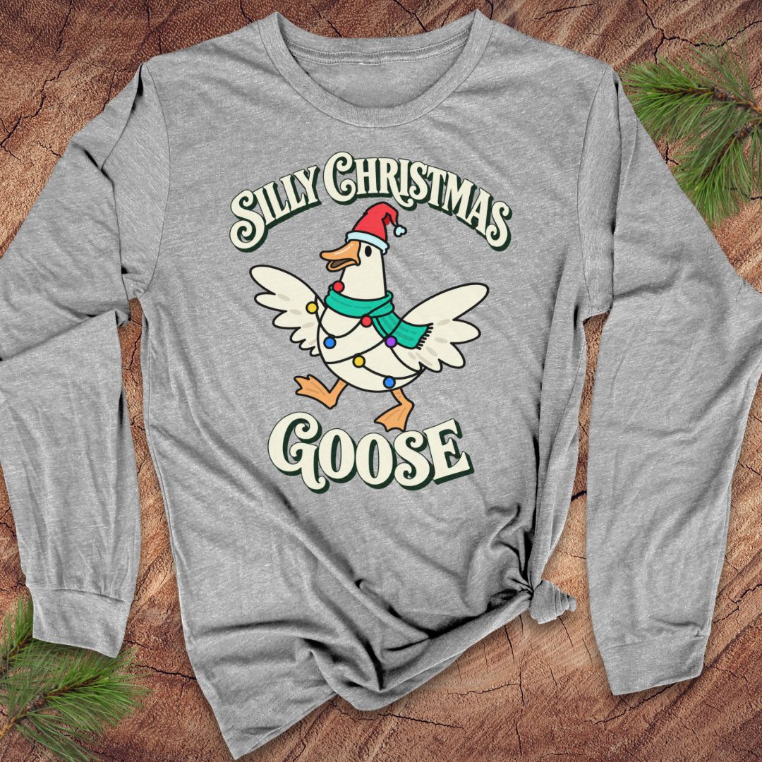 Athletic heather gray long-sleeve t-shirt with a goose in a Santa hat and teal scarf, wrapped in colorful Christmas lights under retro Silly Christmas Goose lettering, laid flat on a rustic wood background with pine branches.