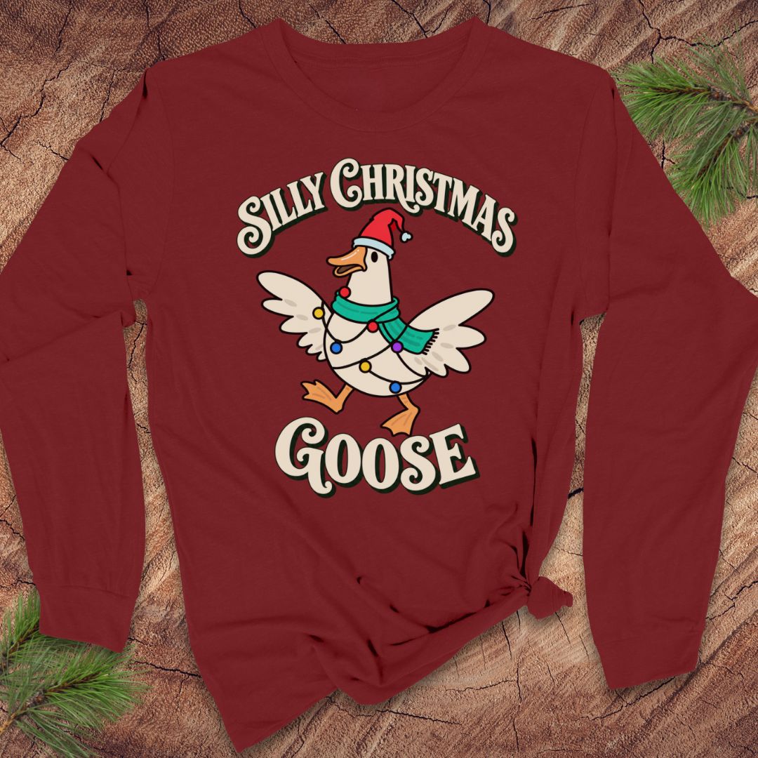 Cardinal red long-sleeve t-shirt with a goose in a Santa hat and teal scarf, wrapped in colorful Christmas lights under retro Silly Christmas Goose lettering, laid flat on a rustic wood background with pine branches.
