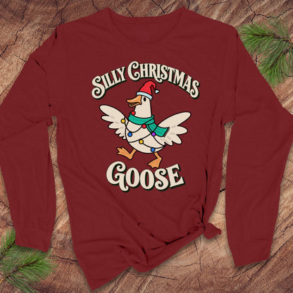 Cardinal red long-sleeve t-shirt with a goose in a Santa hat and teal scarf, wrapped in colorful Christmas lights under retro Silly Christmas Goose lettering, laid flat on a rustic wood background with pine branches.