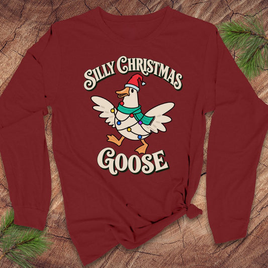 Cardinal red long-sleeve t-shirt with a goose in a Santa hat and teal scarf, wrapped in colorful Christmas lights under retro Silly Christmas Goose lettering, laid flat on a rustic wood background with pine branches.
