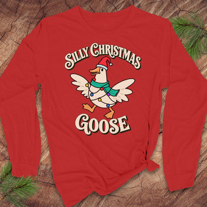 Red long-sleeve t-shirt with a goose in a Santa hat and teal scarf, wrapped in colorful Christmas lights under retro Silly Christmas Goose lettering, laid flat on a rustic wood background with pine branches.