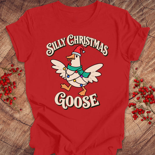 Red t-shirt with a goose in a Santa hat and teal scarf, wrapped in colorful Christmas lights under retro Silly Christmas Goose lettering, laid flat on a rustic wood background with red winter berries.