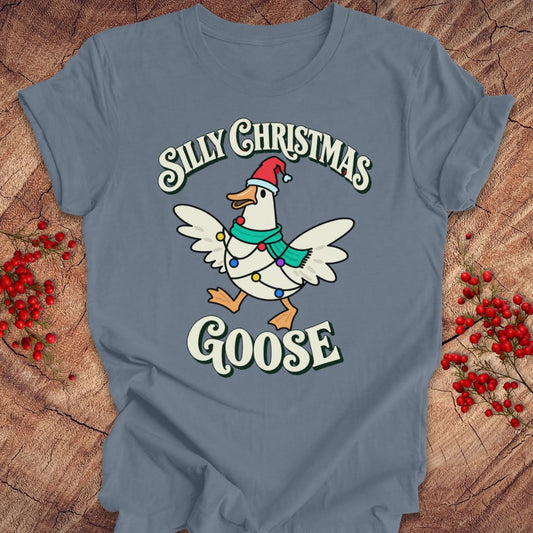 Steel blue t-shirt with a goose in a Santa hat and teal scarf, wrapped in colorful Christmas lights under retro Silly Christmas Goose lettering, laid flat on a rustic wood background with red winter berries.