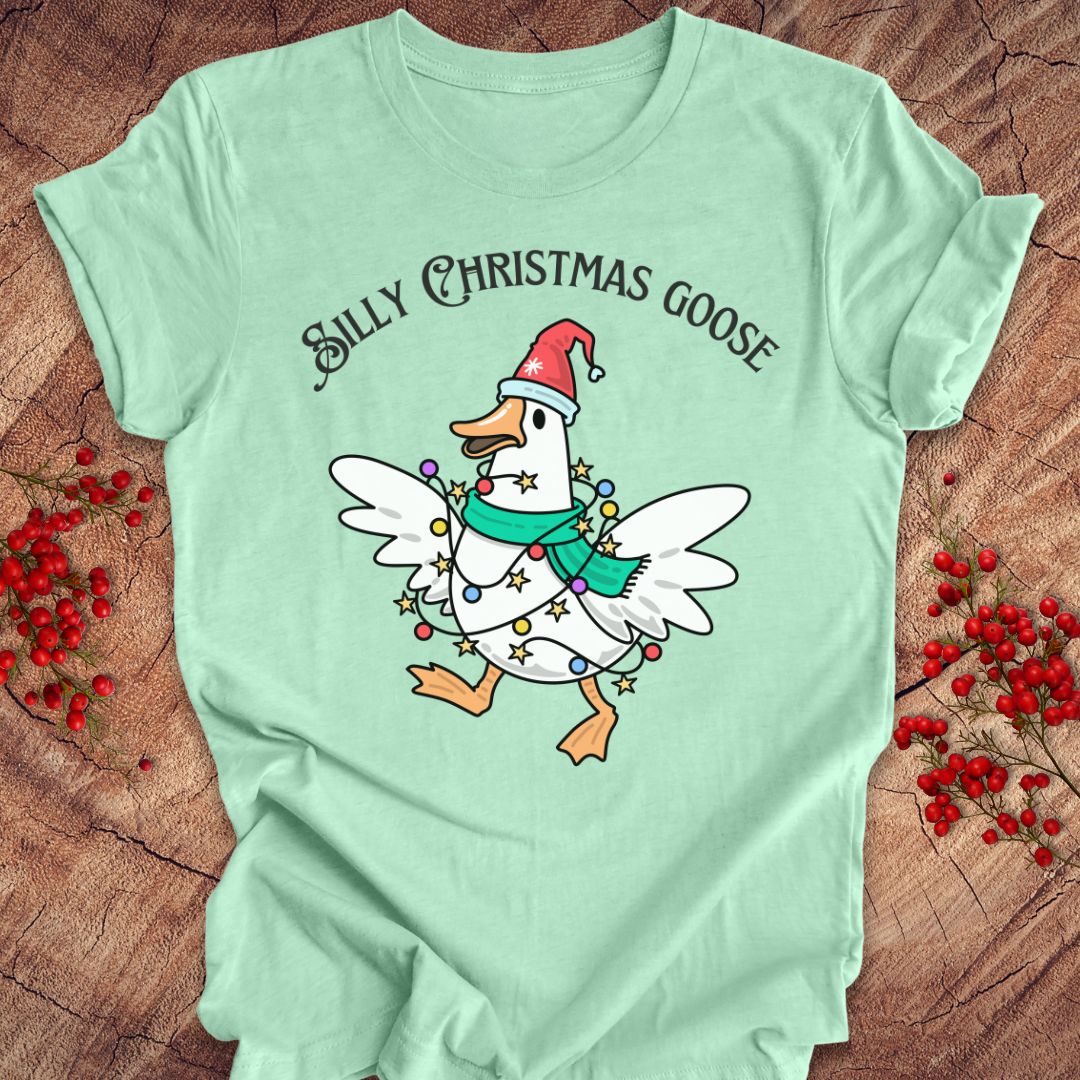 A mint green graphic t-shirt laid flat on a rustic wood background with red berries. The shirt features a cartoon white goose wearing a red Santa hat and a green scarf, decorated with Christmas lights, under the text 'SILLY CHRISTMAS GOOSE'.