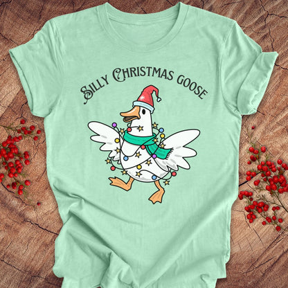 A mint green graphic t-shirt laid flat on a rustic wood background with red berries. The shirt features a cartoon white goose wearing a red Santa hat and a green scarf, decorated with Christmas lights, under the text 'SILLY CHRISTMAS GOOSE'.