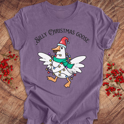 A purple short-sleeve t-shirt laid flat on a rustic wood surface, featuring a cartoon white goose wearing a red Santa hat and green scarf with holiday lights, and the text 'SILLY CHRISTMAS GOOSE' above it. Red berry sprigs are placed around the shirt.
