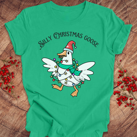 A green short-sleeve T-shirt laid flat on a rustic wood surface, featuring a cartoon Christmas goose wearing a red Santa hat and green scarf with colorful lights, plus the text 'SILLY CHRISTMAS GOOSE' above it. Red berry sprigs are placed around the shirt as styling props.