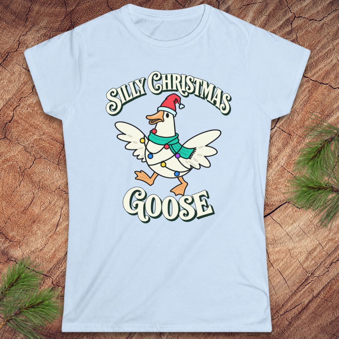 Light blue women's t-shirt with a goose in a Santa hat and teal scarf, wrapped in colorful Christmas lights under retro Silly Christmas Goose lettering, laid flat on a rustic wood background with pine branches.