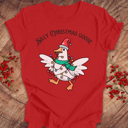 A red short-sleeve graphic T-shirt laid flat on a rustic wood surface with small red berry sprigs around it. The shirt features a cartoon white goose wearing a Santa hat, green scarf, and holiday lights, with the text 'Silly Christmas Goose' above it.