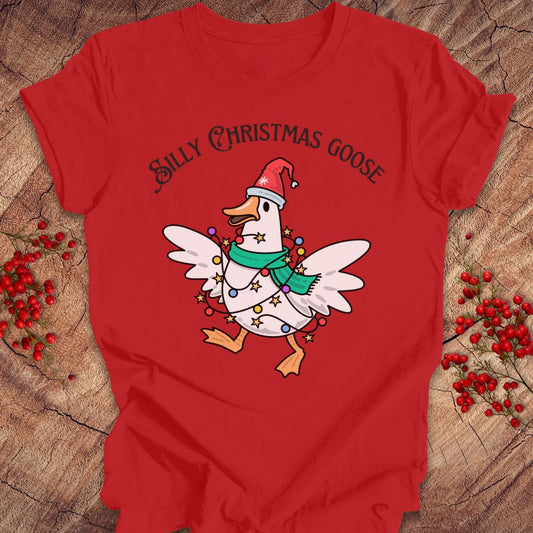 A red short-sleeve graphic T-shirt laid flat on a rustic wood surface with small red berry sprigs around it. The shirt features a cartoon white goose wearing a Santa hat, green scarf, and holiday lights, with the text 'Silly Christmas Goose' above it.