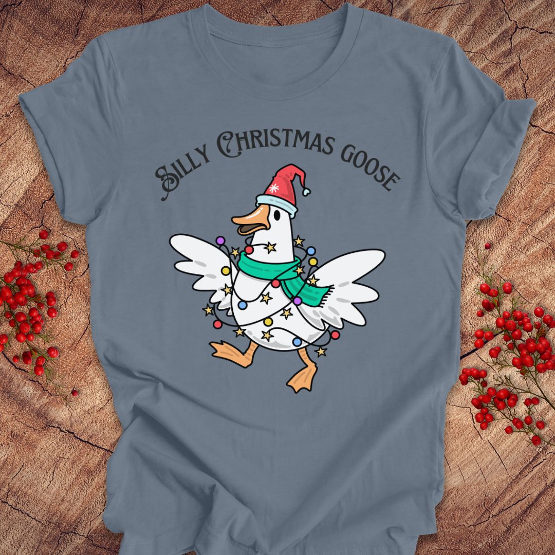 A slate blue graphic t-shirt laid flat on a rustic wood background with red berries. The shirt features a cartoon white Christmas goose wearing a red Santa hat and a green scarf, wrapped in holiday lights, with the text 'SILLY CHRISTMAS GOOSE' above it.