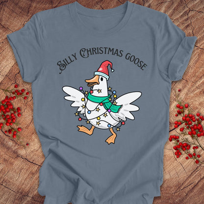 A slate blue graphic t-shirt laid flat on a rustic wood background with red berries. The shirt features a cartoon white Christmas goose wearing a red Santa hat and a green scarf, wrapped in holiday lights, with the text 'SILLY CHRISTMAS GOOSE' above it.