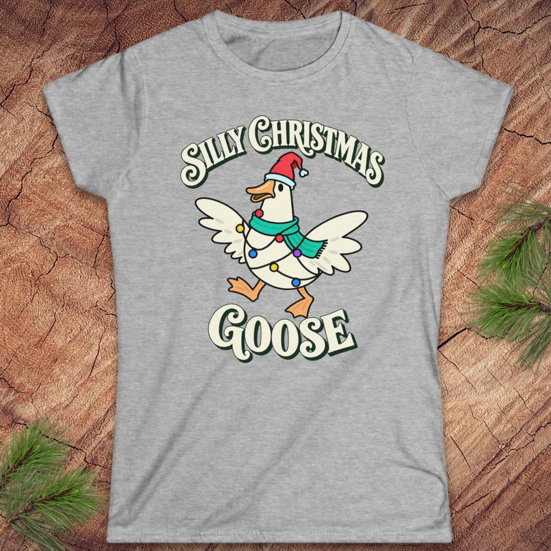 Sport grey women's t-shirt with a goose in a Santa hat and teal scarf, wrapped in colorful Christmas lights under retro Silly Christmas Goose lettering, laid flat on a rustic wood background with pine branches.