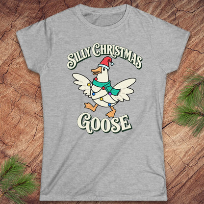 Sport grey women's t-shirt with a goose in a Santa hat and teal scarf, wrapped in colorful Christmas lights under retro Silly Christmas Goose lettering, laid flat on a rustic wood background with pine branches.