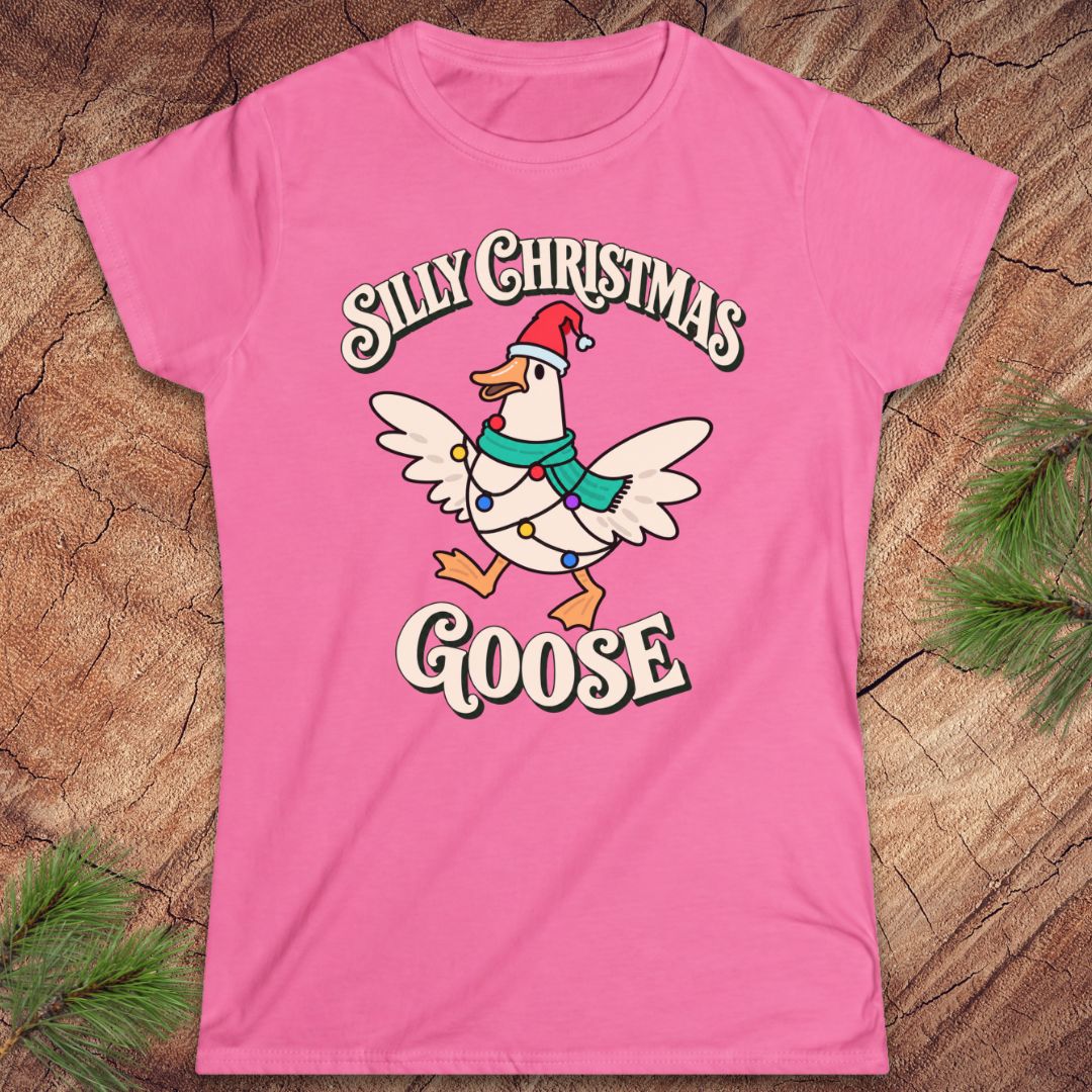 Azalea pink women's t-shirt with a goose in a Santa hat and teal scarf, wrapped in colorful Christmas lights under retro Silly Christmas Goose lettering, laid flat on a rustic wood background with pine branches.