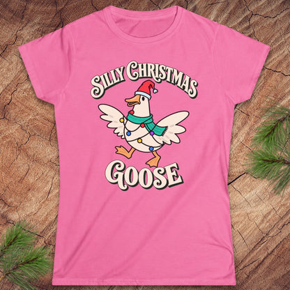 Azalea pink women's t-shirt with a goose in a Santa hat and teal scarf, wrapped in colorful Christmas lights under retro Silly Christmas Goose lettering, laid flat on a rustic wood background with pine branches.